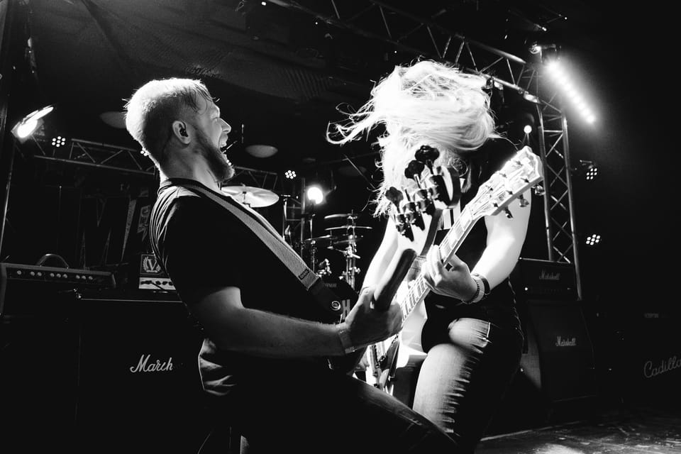 Zwei Gitarristen einer Rockband in voller Bewegung auf der Bühne, die Haare der Bassistin fliegen im Scheinwerferlicht, Schwarzweiß.