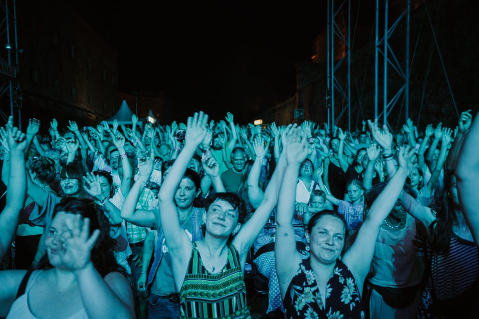 Festival-Publikum bei Nacht in türkisem Bühnenlicht, dutzende Arme in die Höhe gestreckt, vorne zwei lachende Frauen.