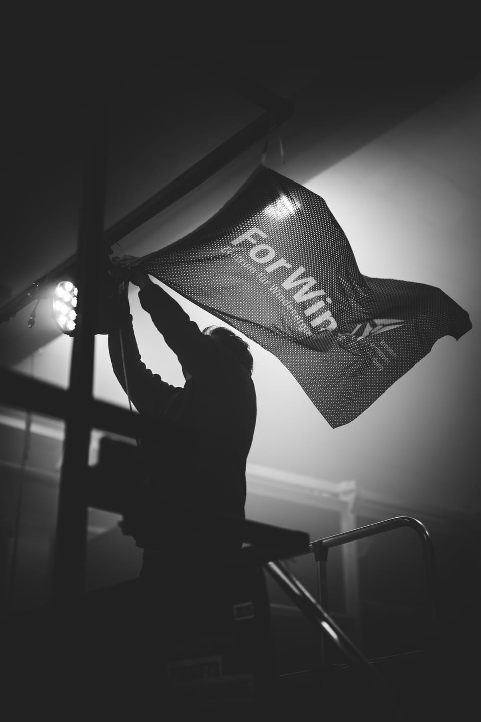 Silhouette eines Mannes, der auf einer Bühnenkonstruktion eine große Fahne schwenkt, von hinten angestrahlt, Schwarzweiß.
