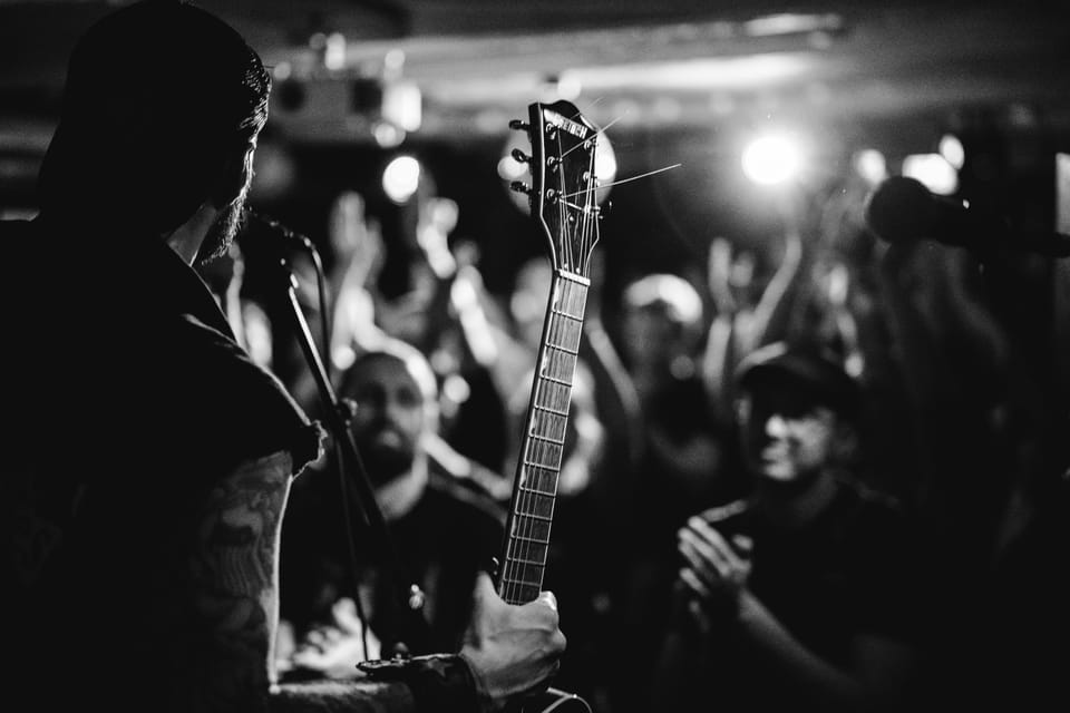 Über die Schulter eines Gitarristen fotografiert: der Gitarrenhals im Vordergrund, dahinter ein Club voller erhobener Hände, Schwarzweiß.