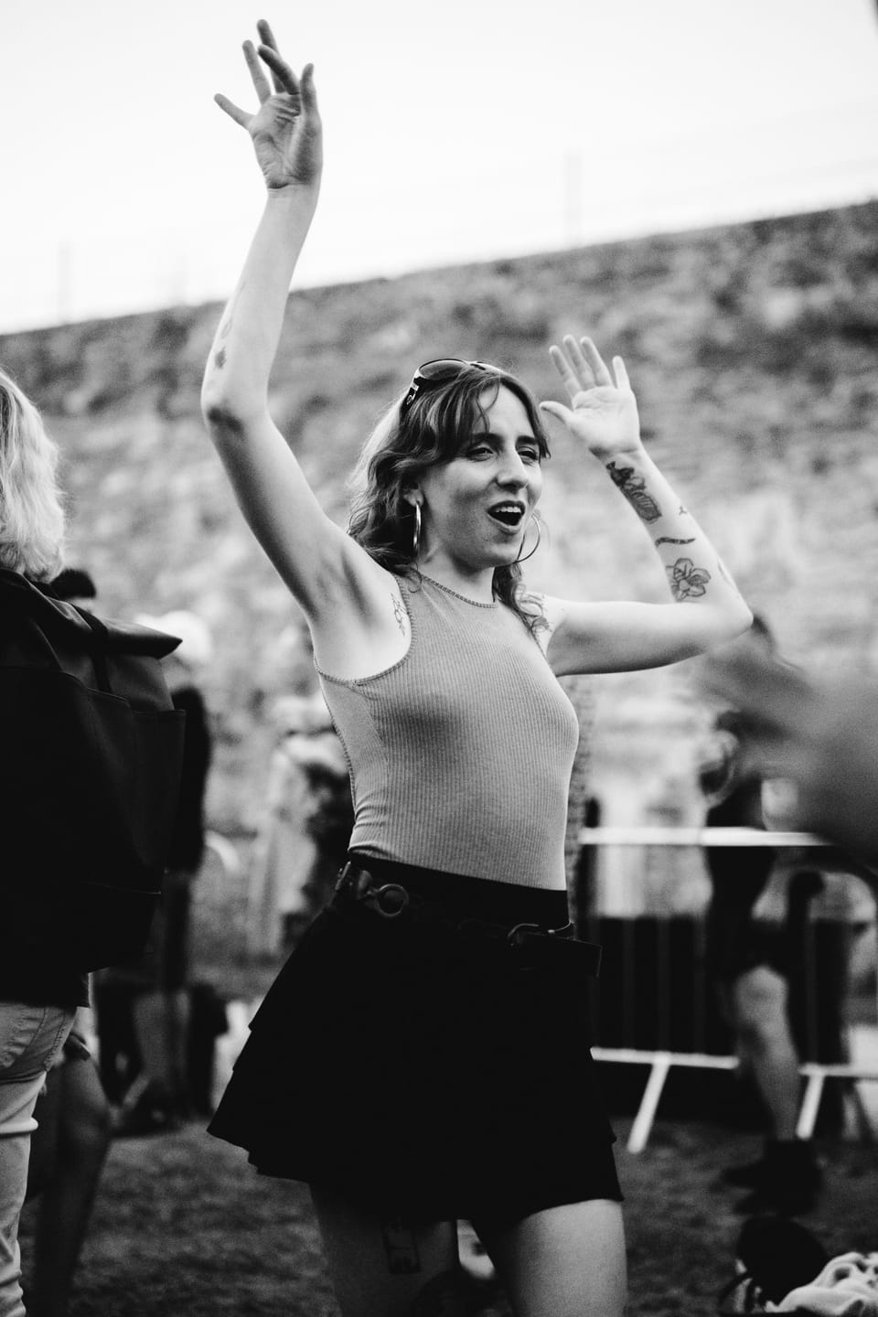 Junge Frau mit Ohrringen und Tattoos tanzt mit beiden Armen über dem Kopf vor einer alten Festivalmauer, Schwarzweiß.