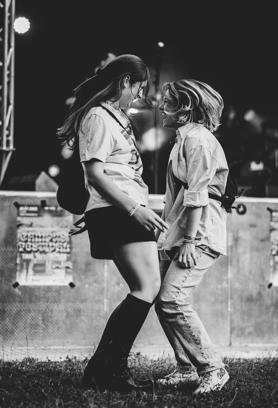 Zwei junge Frauen tanzen sich lachend gegenüber auf einem Festivalgelände, Schwarzweiß, das Haar fliegt.