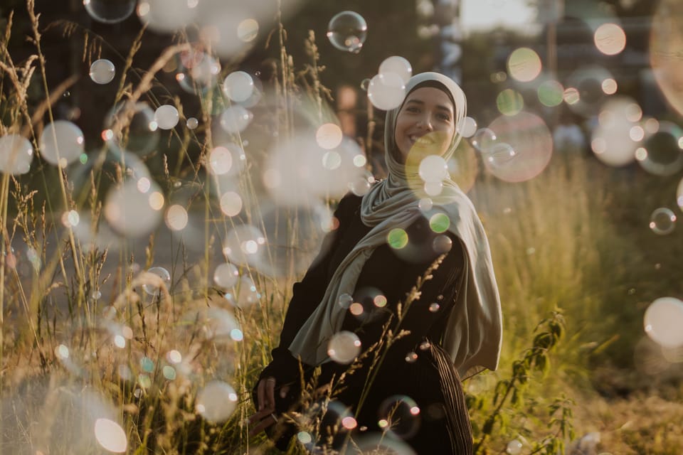 Lächelnde Frau mit hellem Hijab steht in einer sommerlichen Wiese im Abendlicht, umgeben von schwebenden Seifenblasen.