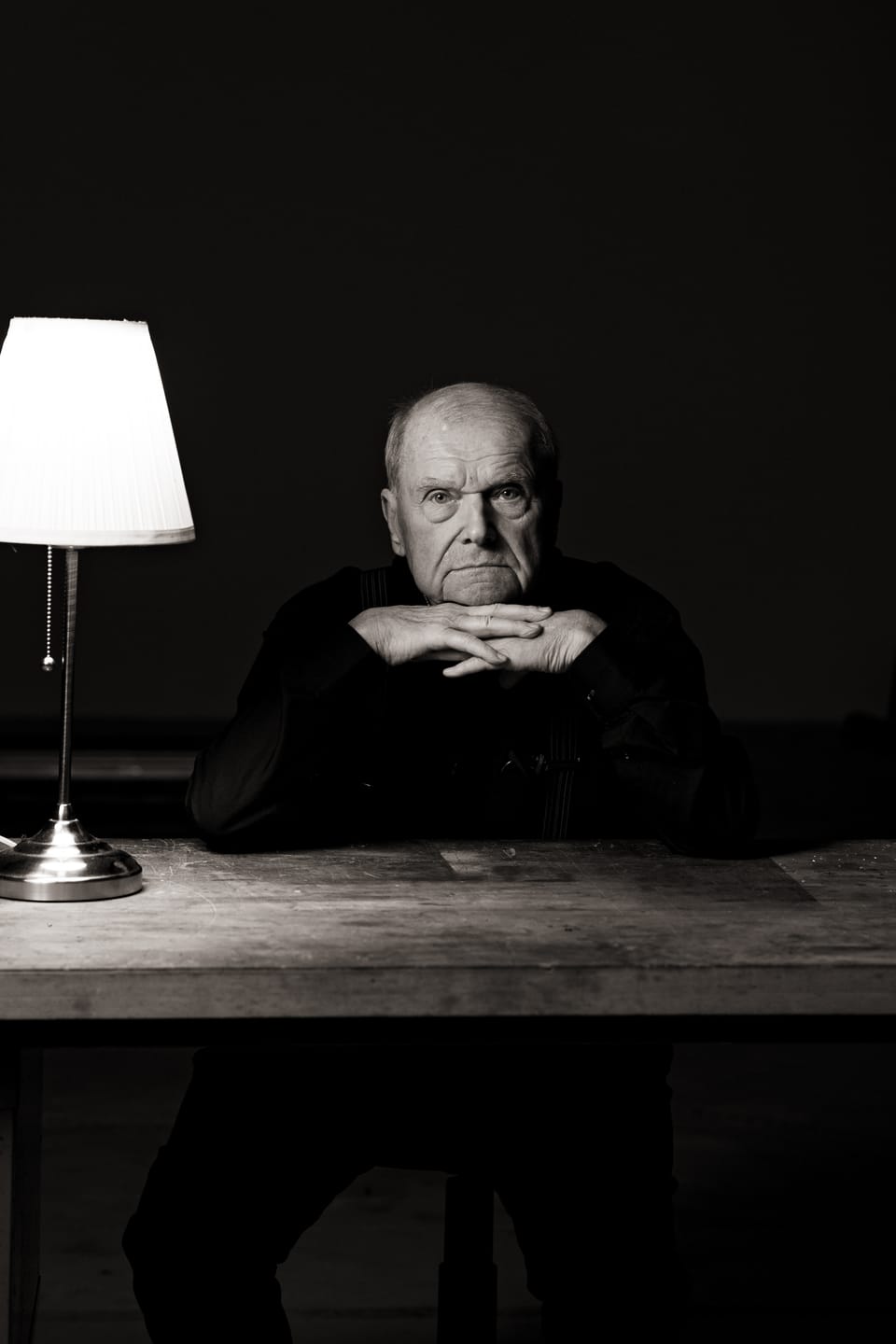 Älterer Herr sitzt ernst an einem Holztisch, das Kinn auf die gefalteten Hände gestützt, neben einer leuchtenden Tischlampe, Schwarzweiß.