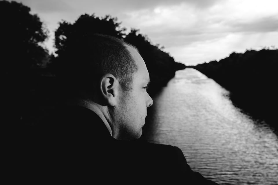 Profil eines Mannes, der von einer Brücke über einen ruhigen Kanal in den dramatischen Abendhimmel blickt, Schwarzweiß.