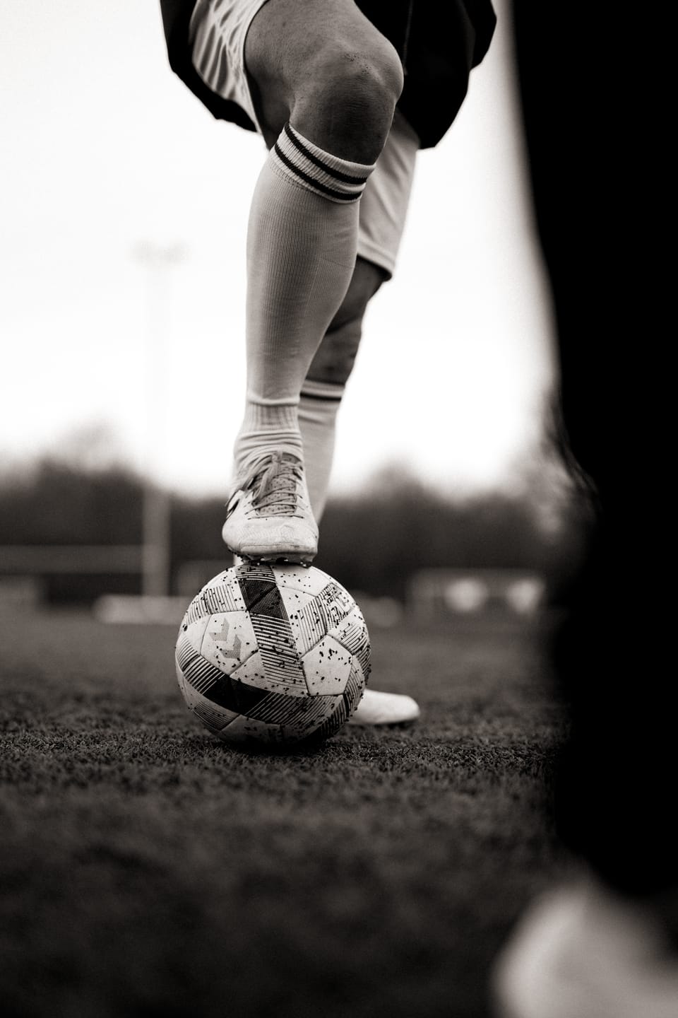 Nahaufnahme in Sepia: ein Fuß mit Stutzen ruht auf einem abgewetzten Fußball auf dem Rasen.