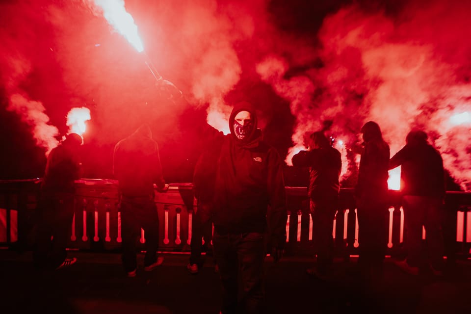 Fangruppe auf einer Brücke bei Nacht, rote Bengalos tauchen Rauch und Silhouetten in grelles Rot.