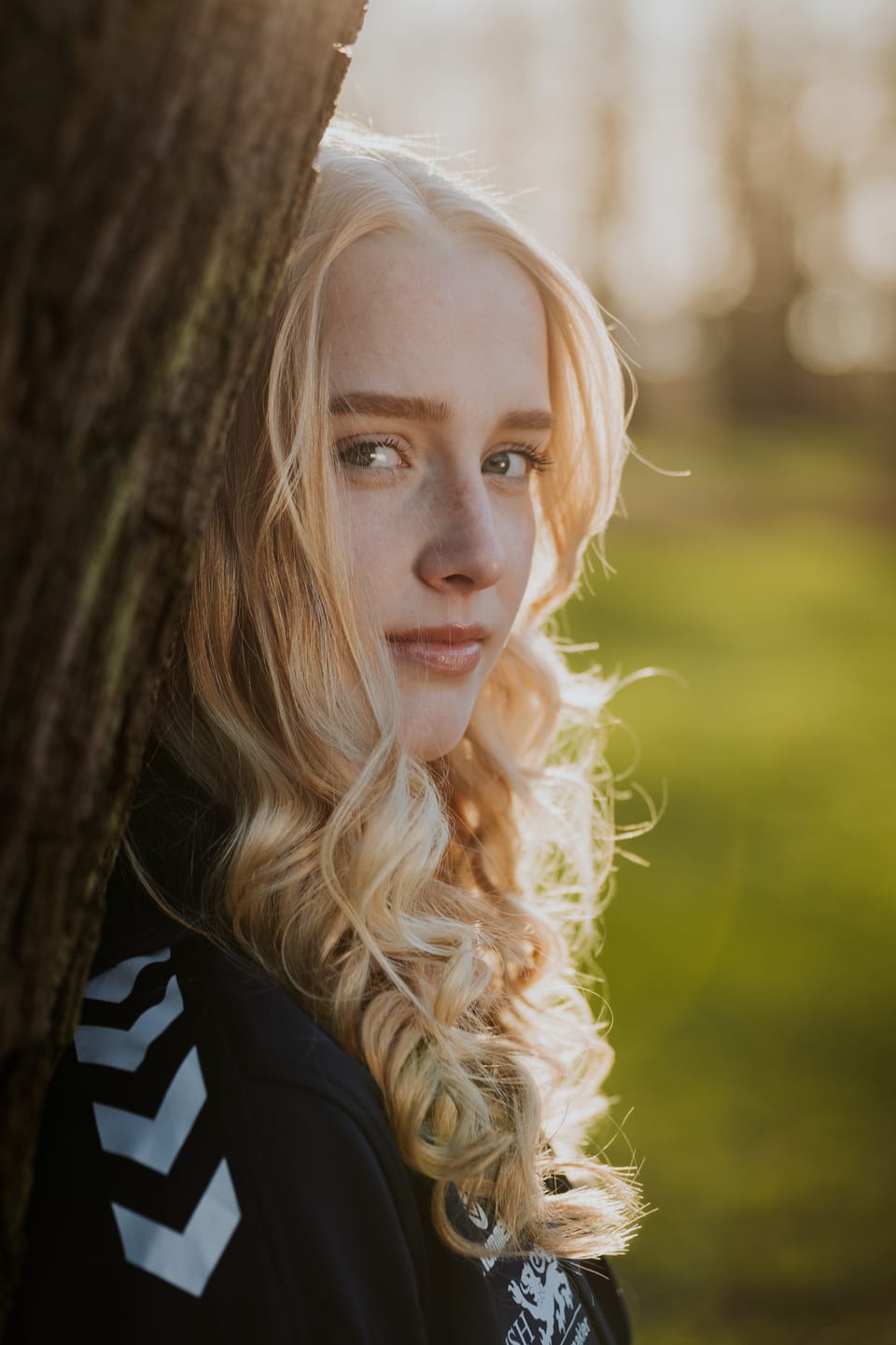 Junge Fußballerin mit langem blondem Haar lehnt im warmen Abendlicht an einem Baum und blickt ruhig in die Kamera.