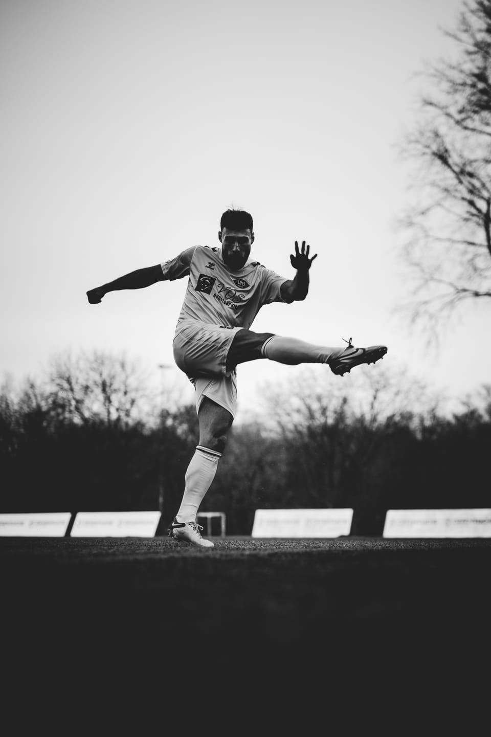 Fußballer im Moment des Volleyschusses, das Bein hoch in der Luft, als dunkle Silhouette gegen den weiten Himmel, Schwarzweiß.